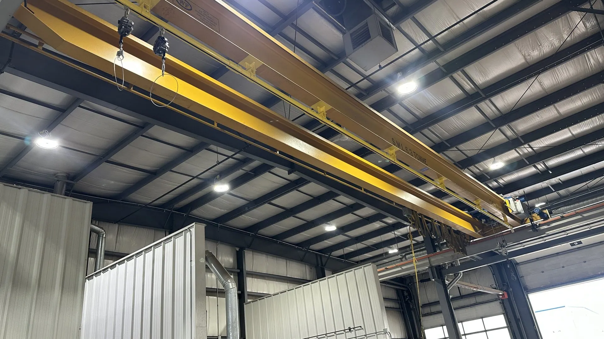 A 44 ft yellow rigid rail fall arrest system mounted beneath the bridge of a 6.3 tonne overhead crane in Alberta, with two self-retracting lifelines hanging from trolleys.