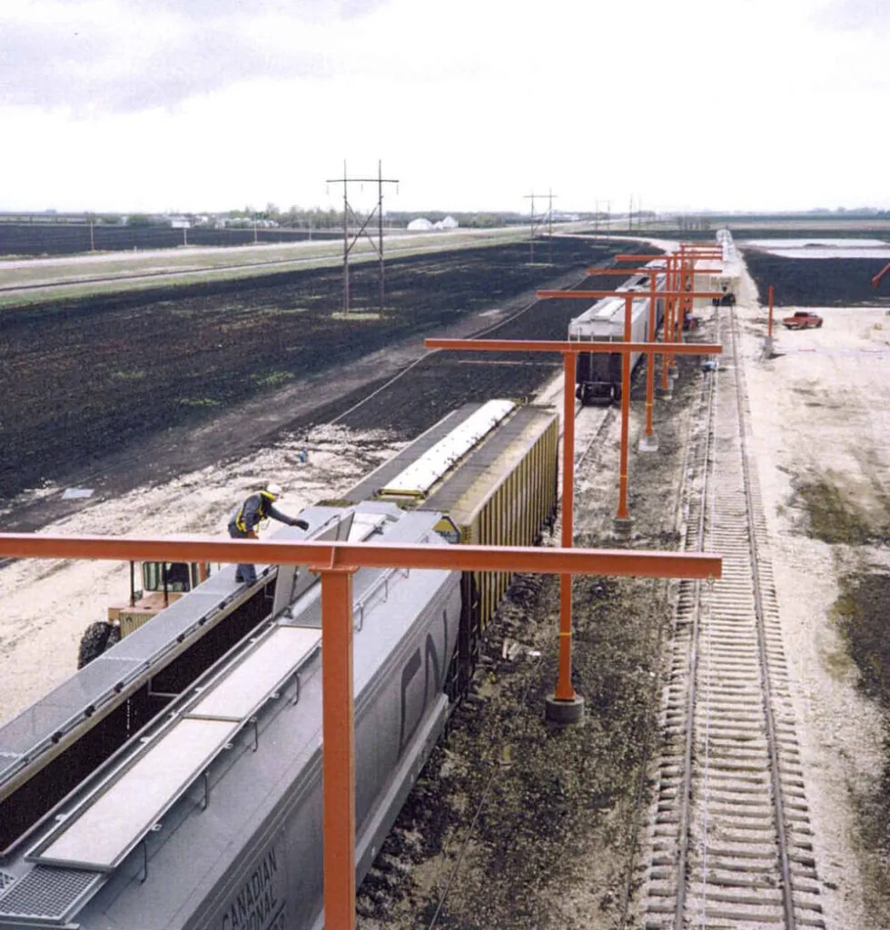 Engineered horizontal lifeline on orange stanchions above hopper cars at a railcar loading system, worker tied off on the car top