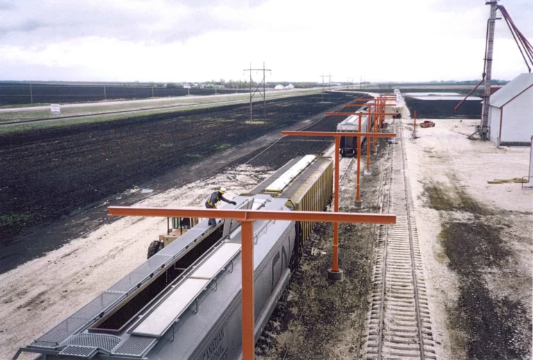 Orange T-frame overhead rail posts over hopper railcars along a loading track