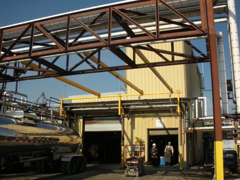 Yellow overhead rigid rail at a tanker truck loading bay under a steel pipe bridge